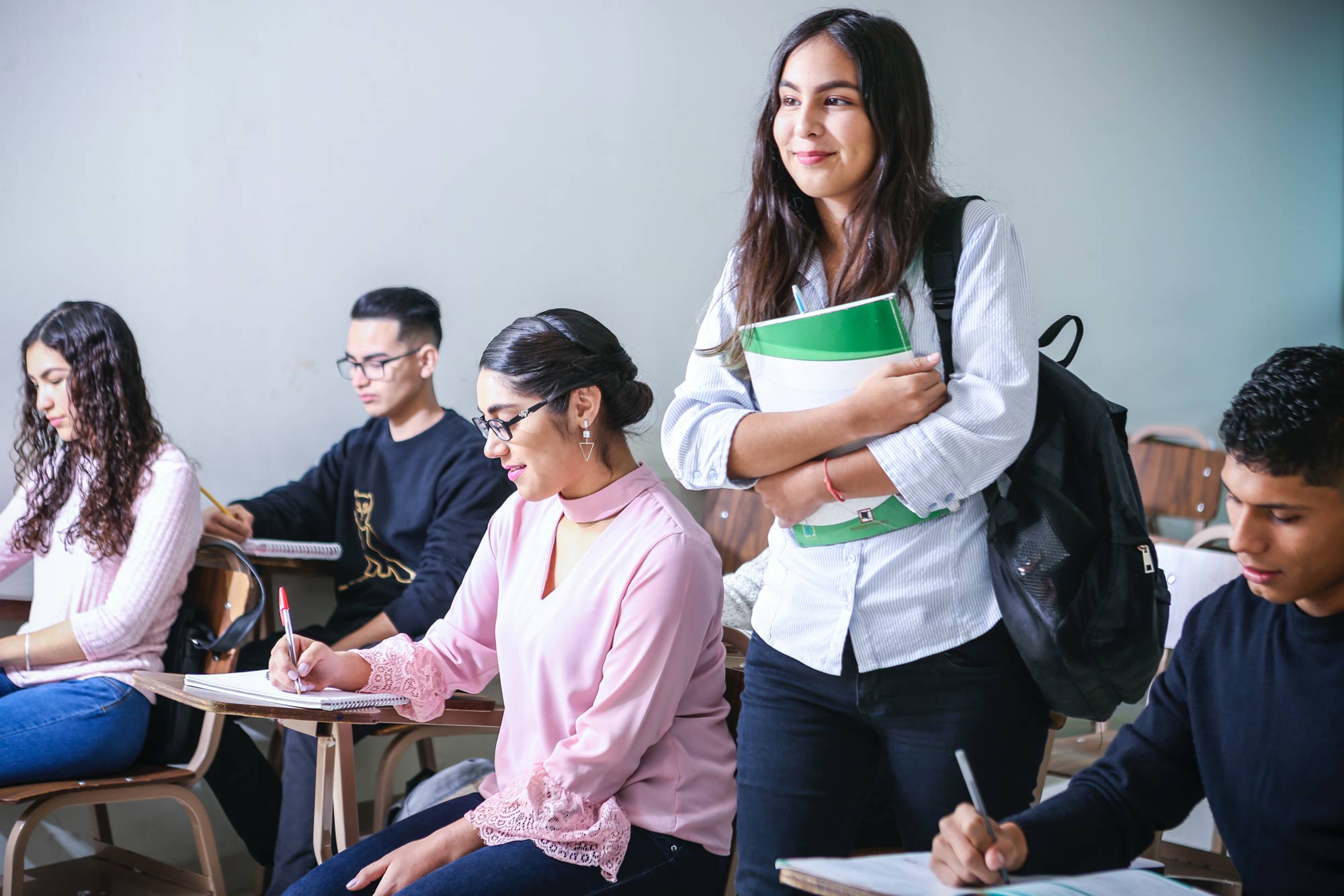 Students at desks in a classroom, one standing with a folder under her arm